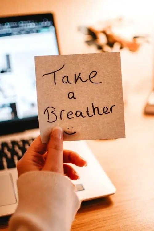 A woman holds a post-it that says "Take a Breather" in front of her computer