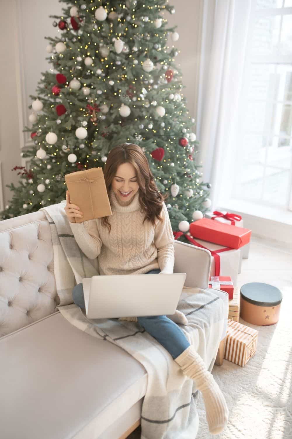 A woman holds a present in front of a computer near a Christmas tree